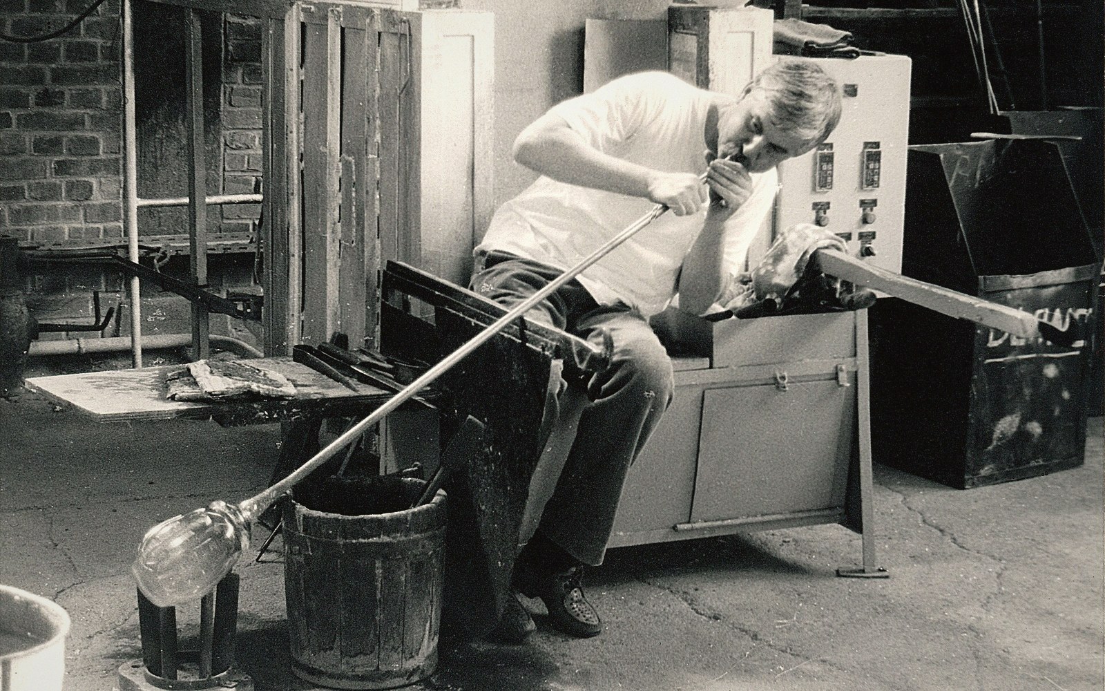 A glassblower sits at a bench, blowing into a long pipe to shape molten glass
